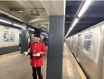 A man wearing a British soldier outfit at 2nd Ave Station walking on the platform between the R179 consist F train and the Arnine consist making up the Holiday Train